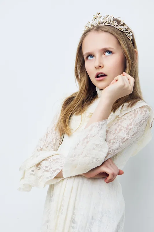 Young girl in a beautiful white dress and crown, pondering thoughtfully against a light background, showcasing innocence and elegance