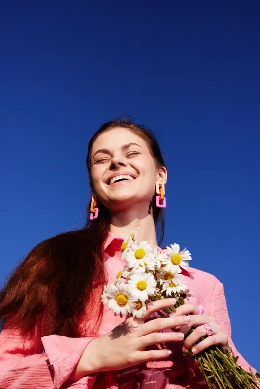 Joyful young woman with daisies under a clear blue sky, wearing a pink shirt and vibrant earrings, embodying happiness and positivity