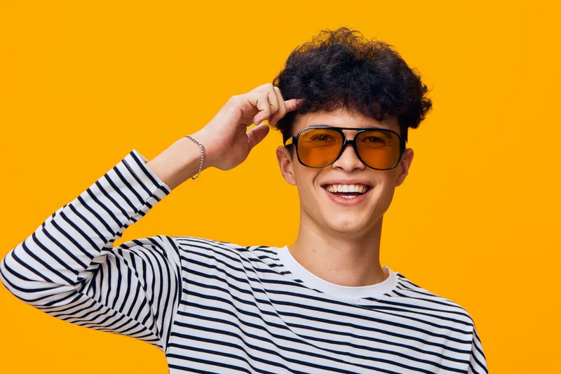 smiling man with sunglasses wearing striped shirt poses happily against bright orange background, showcasing youthful energy and casual style.