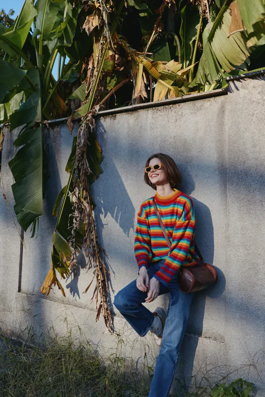Woman in her mid-20s wearing a colorful striped sweater and sunglasses, smiling and leaning against a concrete wall with banana leaves, blue jeans and crossbody bag for tropical lifestyle travel