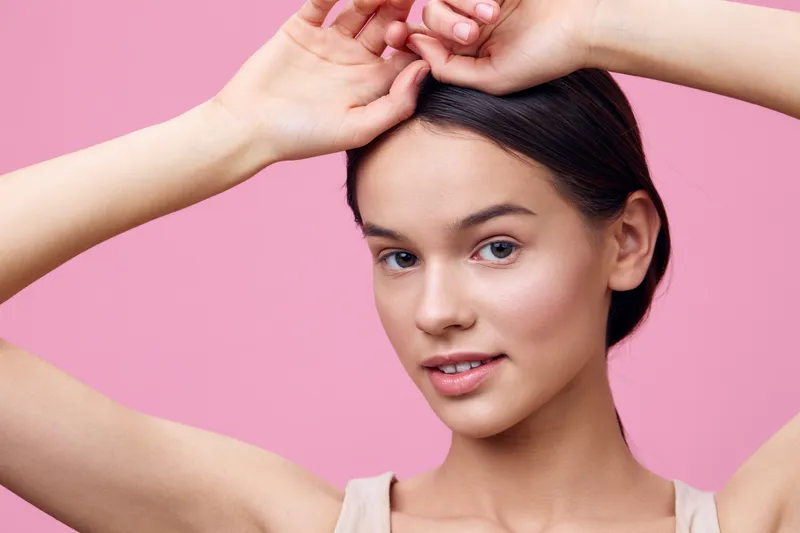 Young woman with clear skin and a natural pose against a pink background, expressing confidence and beauty in a minimalistic style, ideal for beauty and skincare themes