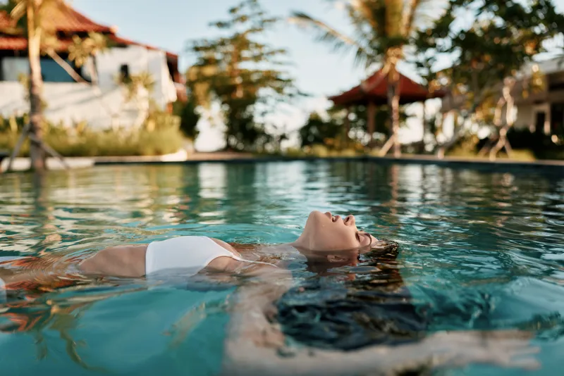 Relaxed woman floating in a sunlit pool, serene water, outdoor retreat, warm breeze, clear blue hues, summer leisure, candid moment, peaceful vibe, natural colors, bright sky
