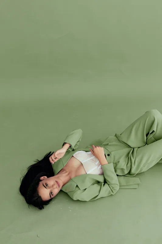 woman lying on green floor wearing stylish green suit and white lace top with relaxed expression in minimalistic studio setting