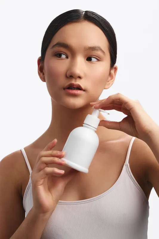 Asian woman holds a small bottle near her face, preparing to apply skincare product. She looks calm and focused, conveying a scene of personal care and wellness in a clean studio setting. The image