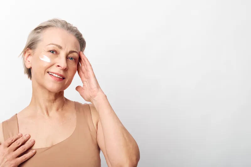 Mature woman with light skin applies moisturizing cream on face, smiling gently. Soft natural lighting highlights healthy skincare routine and self-care concept on neutral background.