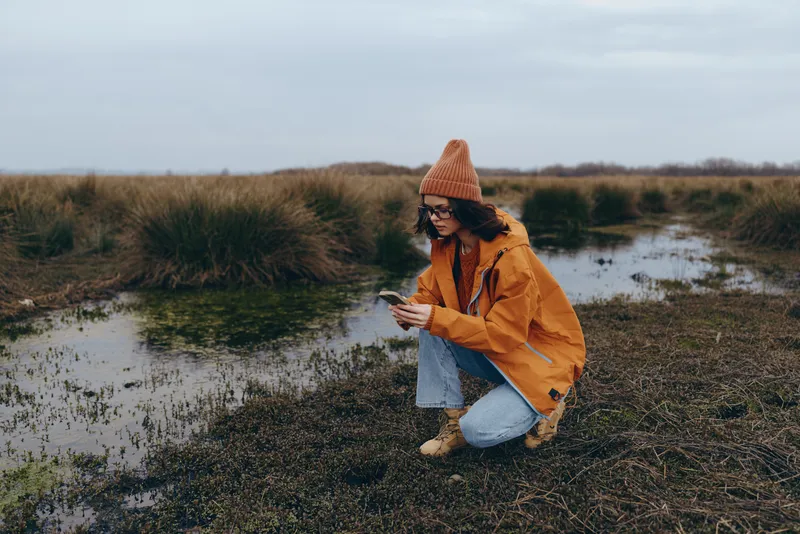 Woman lifestyle outdoors kneeling by water in wetlands, wearing orange coat and knit hat, holding smartphone while crouching, calm autumn nature mood, soft overcast light, countryside travel