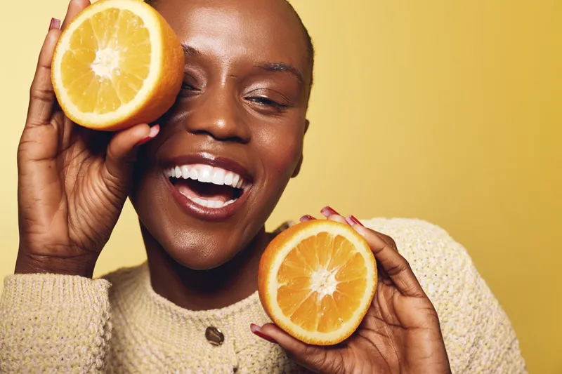 Happy African woman with dark skin holds two halves of fresh orange close to her face on yellow background. Concept of healthy lifestyle, vitamin C, skincare, and positive mood.