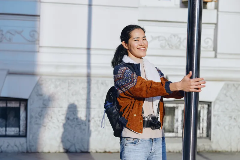 Smiling woman outdoors by a light building, wearing a scarf and jacket, backpack on shoulder, camera hanging, gripping a pole, sun casting shadows along the scene