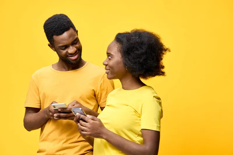 Young African couple smiling at each other while using smartphones, dressed in bright yellow outfits, against a vibrant yellow background, conveying joy and connection