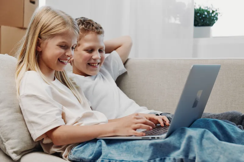 Two smiling children sitting on a couch using a laptop together in a bright room. Concept of learning, technology, friendship, and digital entertainment for kids.