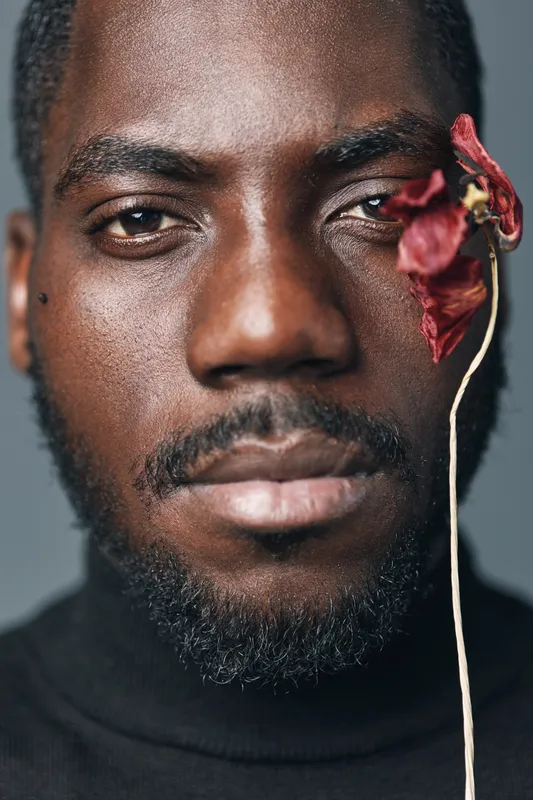 African man portrait with dark skin and beard in creative conceptual style holding dried flower near face. Closeup, studio lighting and artistic expression with serious mood and black turtleneck.