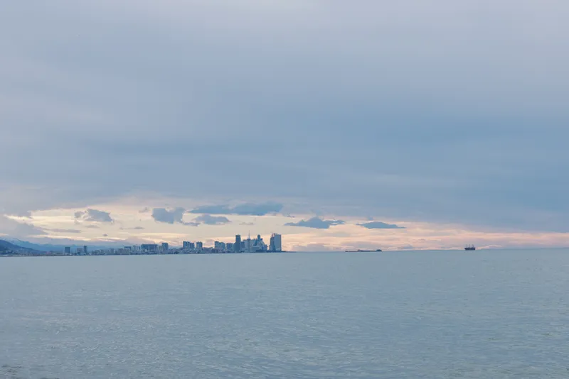 Distant city skyline along a calm seascape under moody clouds, tranquil water and a soft horizon over the ocean at dusk.
