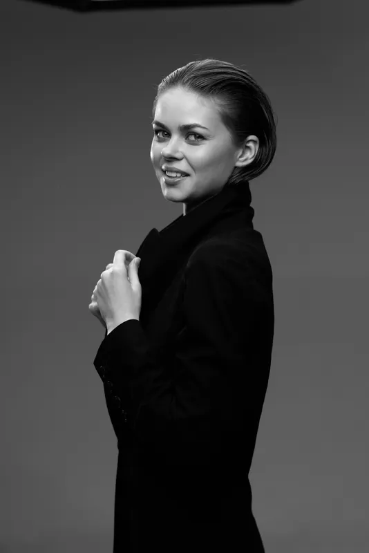 Black and white portrait of smiling woman in black coat with slicked-back hair against neutral background