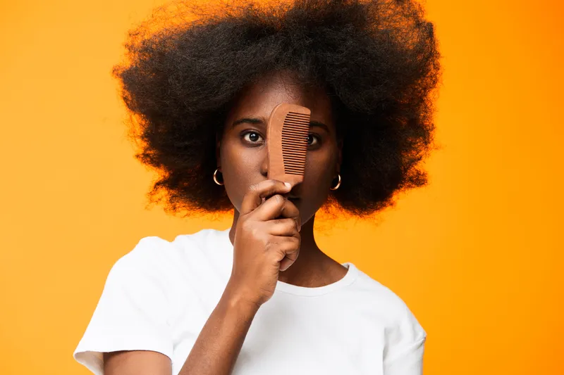 Young woman holding a comb in front of her face, showcasing natural hair texture against a vibrant orange background, evoking confidence and beauty in fashion