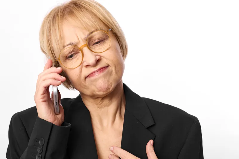 Elderly woman with glasses in a black blazer speaks on a phone call hand at chest and worried thoughtful look isolated on white background communication concept