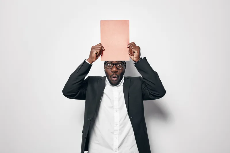 African American man with dark skin wearing glasses and suit holding pink paper above head with surprised expression on white background.