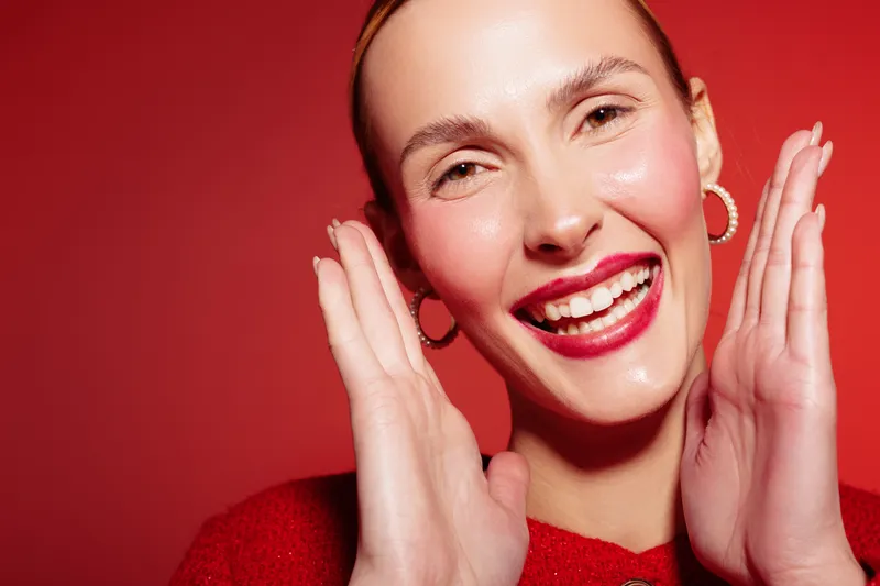 Smiling woman with red lipstick and hoop earrings poses on red background. Close-up portrait with joyful expression and natural skin glow. Concept of happiness and beauty.