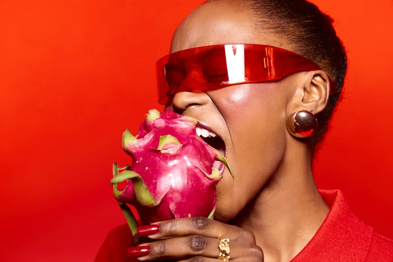 African woman with dark skin wearing red futuristic sunglasses bites a fresh dragon fruit. Bright red background and fashion accessories create a bold and vibrant concept of healthy lifestyle.
