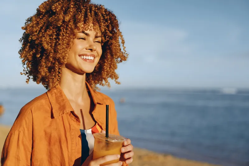 Woman with curly hair smiling outdoors, holding a cold drink, on seaside beach, warm sunlight, golden hour breeze, casual orange jacket, calm ocean horizon