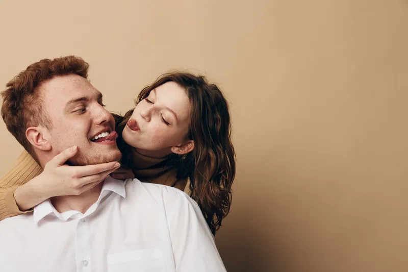 Happy young couple in casual clothes playfully interact with each other on beige background. Woman holds man’s face while he smiles and sticks out his tongue. Concept of love and fun.