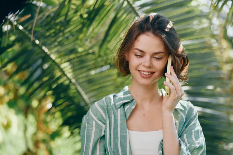 A smiling woman talks on a mobile phone outdoors among lush green tropical plants, wearing a striped green shirt.