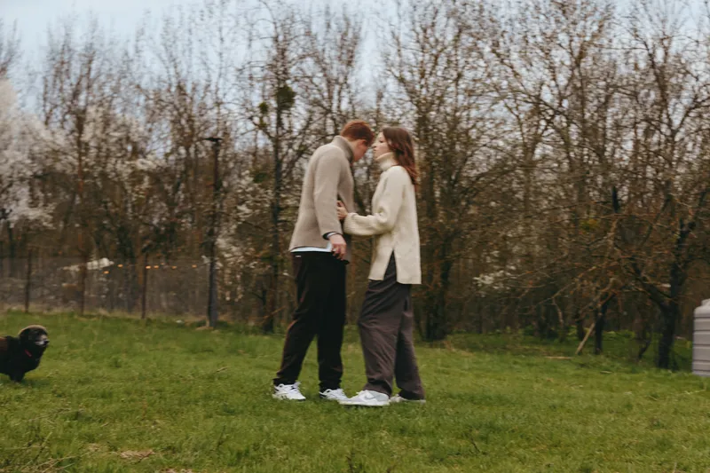Two adults share a tender moment in a sunlit park, embracing while strolling on green grass with bare trees in the background. Casual, cozy mood highlights love, companionship, and outdoor relaxation.