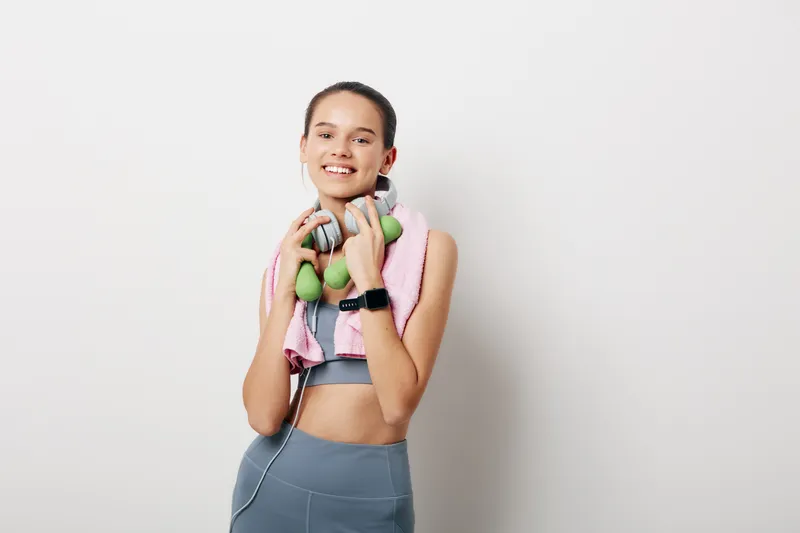 Young woman fitness portrait holding green dumbbells and headphones, wearing gray sportswear and pink towel, smartwatch on wrist, smiling against plain white wall, active lifestyle concept.
