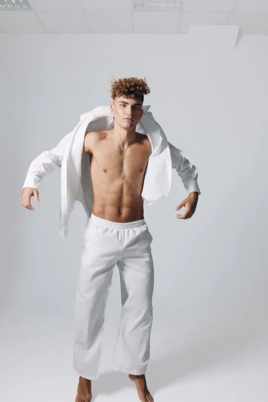 Young man with curly hair wearing white pants and open white shirt standing barefoot in studio with plain background, showing toned body and confident pose for fashion or fitness.