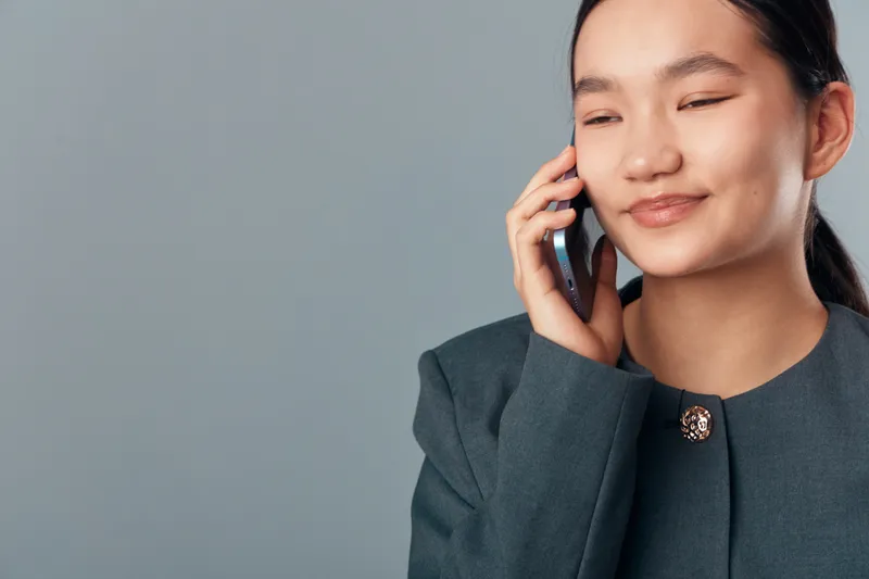Young woman smiling and talking on mobile phone with eyes closed wearing formal jacket for business communication and positive conversation in office environment with gray background.