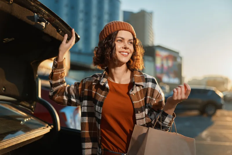 Woman shopping by car trunk in urban parking, smiling with paper bag at golden hour glow. Candid lifestyle portrait conveying authenticity, mindful living and emotional storytelling mood.