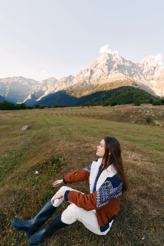 Woman sitting in meadow in autumn with mountains in background, landscape portrait of nature scene. Young female in boots and jacket enjoying serene alpine view.