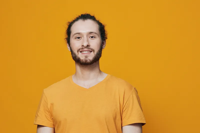 Smiling young man with curly hair wearing an orange t shirt against a vibrant yellow background, embodying happiness and positivity