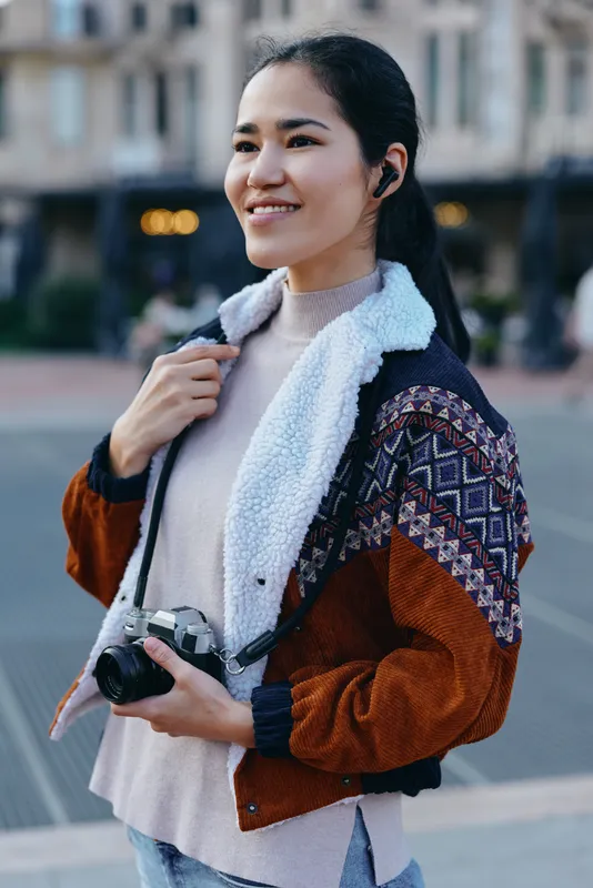 Camera in hand, photographer smiles outdoors on a street, wearing a cozy knitted jacket with shearling collar, ready to capture candid city moments.