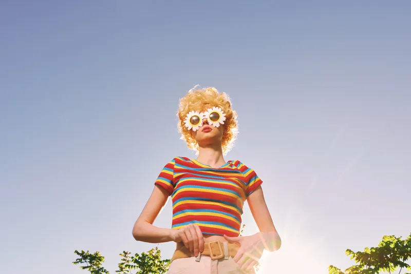 Cheerful woman outdoors, wearing a striped top and sunflower sunglasses, enjoying sunlight, retro film color palette, relaxed pose, natural greenery background, warm breeze, vibrant tones