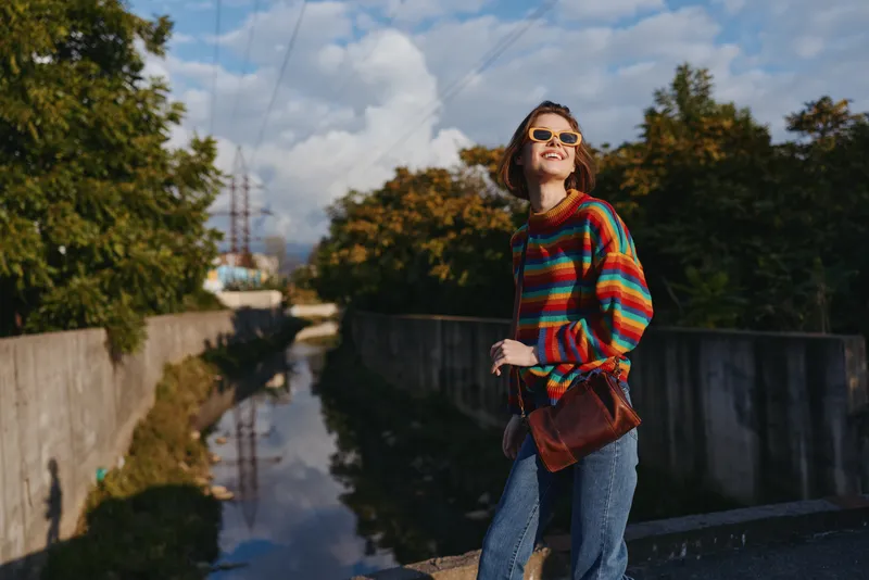 Woman in a colorful striped sweater and sunglasses smiling by an urban canal, mid-20s model in denim jeans with a brown crossbody bag, casual travel lifestyle, confident and sunny autumn walk.