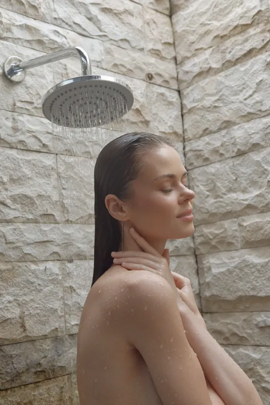Relaxed woman enjoying outdoor shower with stone wall background, clear water droplets, natural light highlighting calm facial expression and wet hair.
