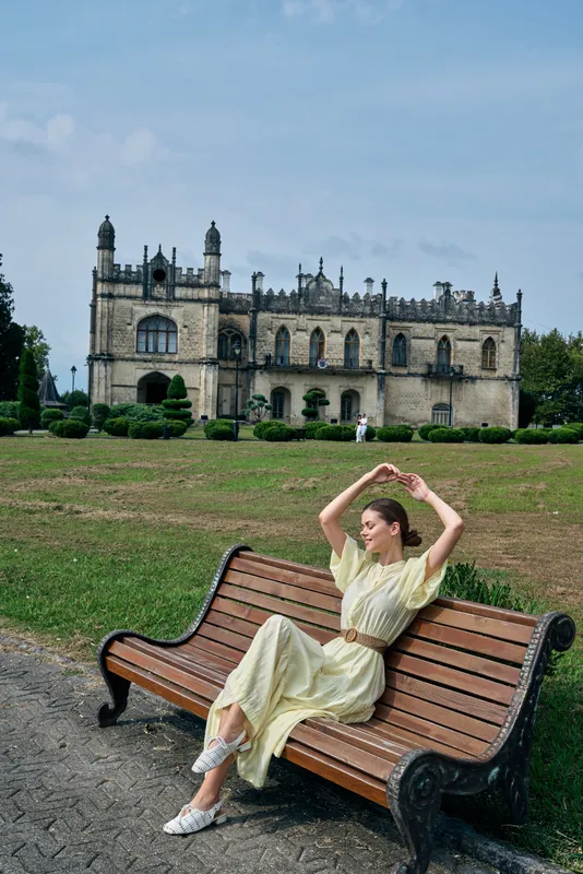 Woman sits on a bench in a historic park garden, enjoying outdoor leisure as a castle looms in the background on a sunny day.