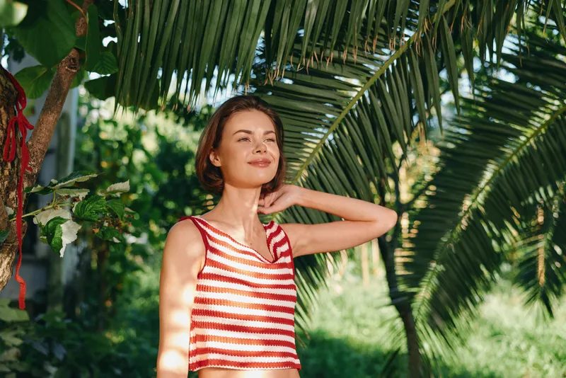 A cheerful woman in a red and white striped sleeveless top stands among lush tropical plants, smiling softly and enjoying a sunny outdoor moment.