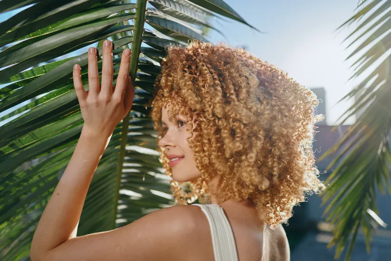 young woman with curly hair gently touching green palm leaves outdoors in bright sunlight, natural beauty, summer mood, relaxed atmosphere
