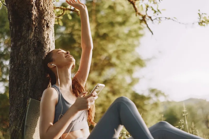 Young woman enjoying music outdoors with a smartphone, expressing joy and relaxation in a sunny park setting, ideal for wellness and lifestyle themes