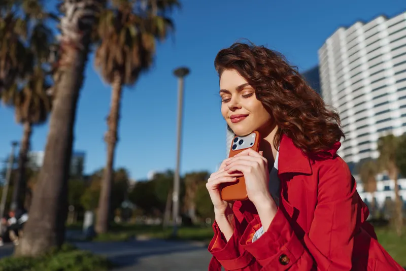 Young woman in a red coat smiles softly while holding a takeaway drink outdoors on a sunny day near palm trees and modern buildings