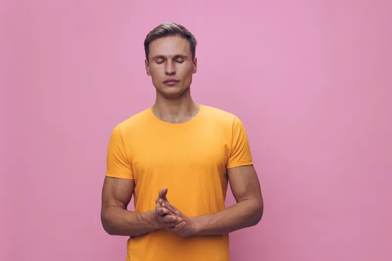 Calm young man with short dark hair and light skin wearing a bright yellow t-shirt stands against a plain pink background, practicing meditation or mindfulness, promoting mental health, relaxation, an