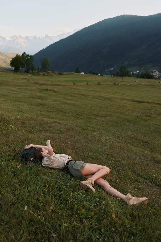 Relaxed woman lying on grass in a wide countryside field with distant mountains, capturing a casual outdoor lifestyle moment of rest, freedom, and connection with nature.