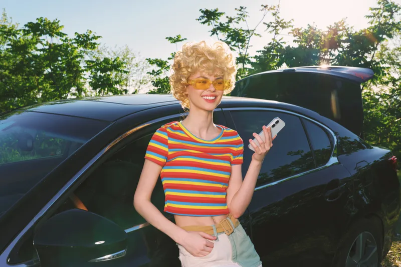 Smiling woman with curly hair wearing a striped croptop stands beside a sleek car on a sunny day, expressing a vibrant lifestyle and retro fashion mood. Soft daylight enhances warm tones, suggesting