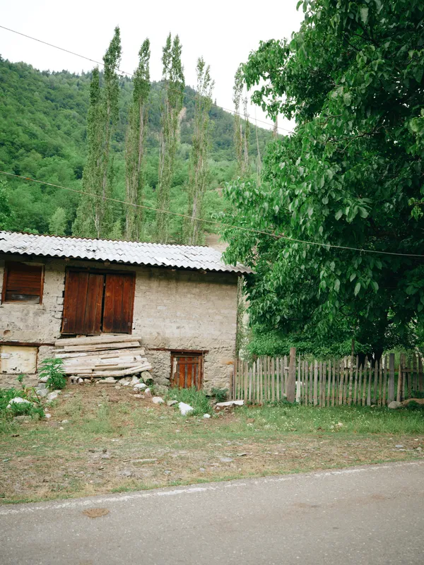 Rural house in a quiet countryside setting with a simple stone facade, wooden shutters, a small yard, and a wooden fence beside a paved road, surrounded by green trees and distant hills.