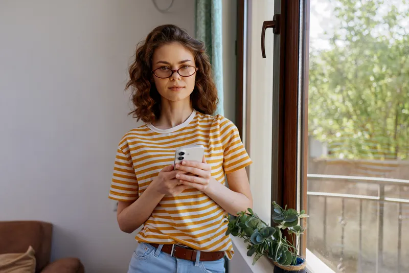Young woman with curly hair and glasses stands by a sunlit window in a casual home setting, scrolling on her smartphone with a relaxed and focused expression.
