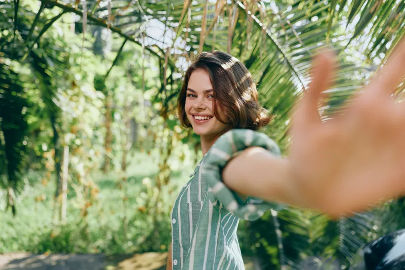 Young woman outdoors, smiling and reaching toward the camera, bathed in bright natural light with a lush garden backdrop and casual striped shirt.
