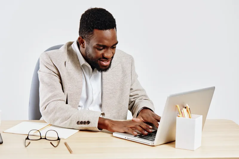 Smiling young African American man working on a laptop at a wooden desk, with notebooks and glasses nearby, in a bright modern office interior. Man focused and engaged, dressed in smart casual attire.