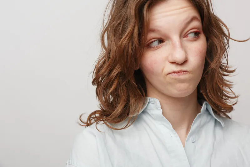 Young woman with curly brown hair looks sideways with skeptical expression wearing white shirt. Isolated on white background. Concept of doubt, suspicion, and uncertainty in emotions.