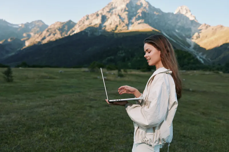 A cheerful outdoor professional smiles while using a laptop in a scenic meadow with mountains in the background, conveying focus, innovation, and balance in a natural work setting.